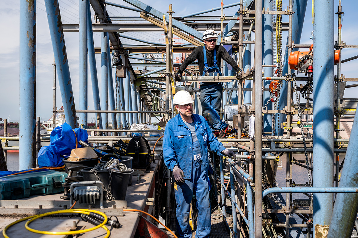 Zwei Industriearbeiter mit Schutzhelmen arbeiten auf einem Gerüst an einer großen Stahlkonstruktion über dem Wasser