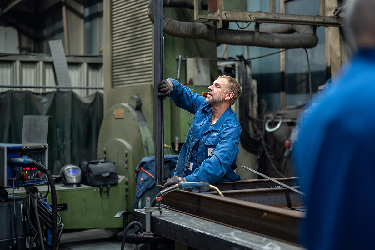 Mitarbeiter in blauer Arbeitskleidung richtet ein Metallgestell in der Werkhalle aus
