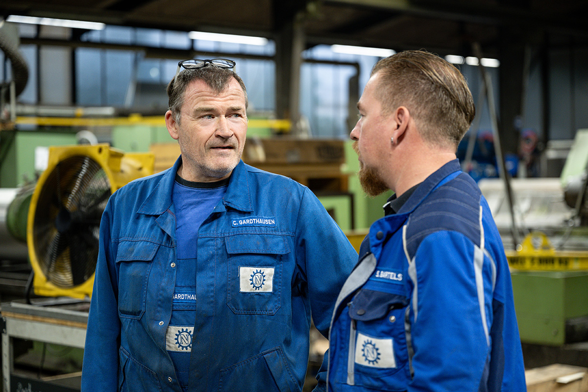 Zwei Mitarbeiter in blauer Arbeitskleidung im Gespräch in der Werkhalle
