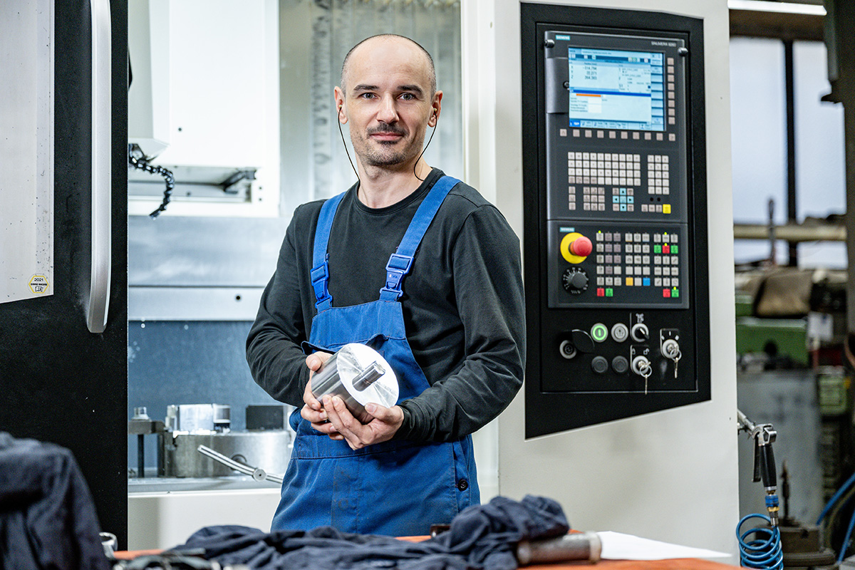 Mitarbeiter in blauer Arbeitskleidung hält ein präzise gefertigtes Metallteil vor einer CNC-Maschine