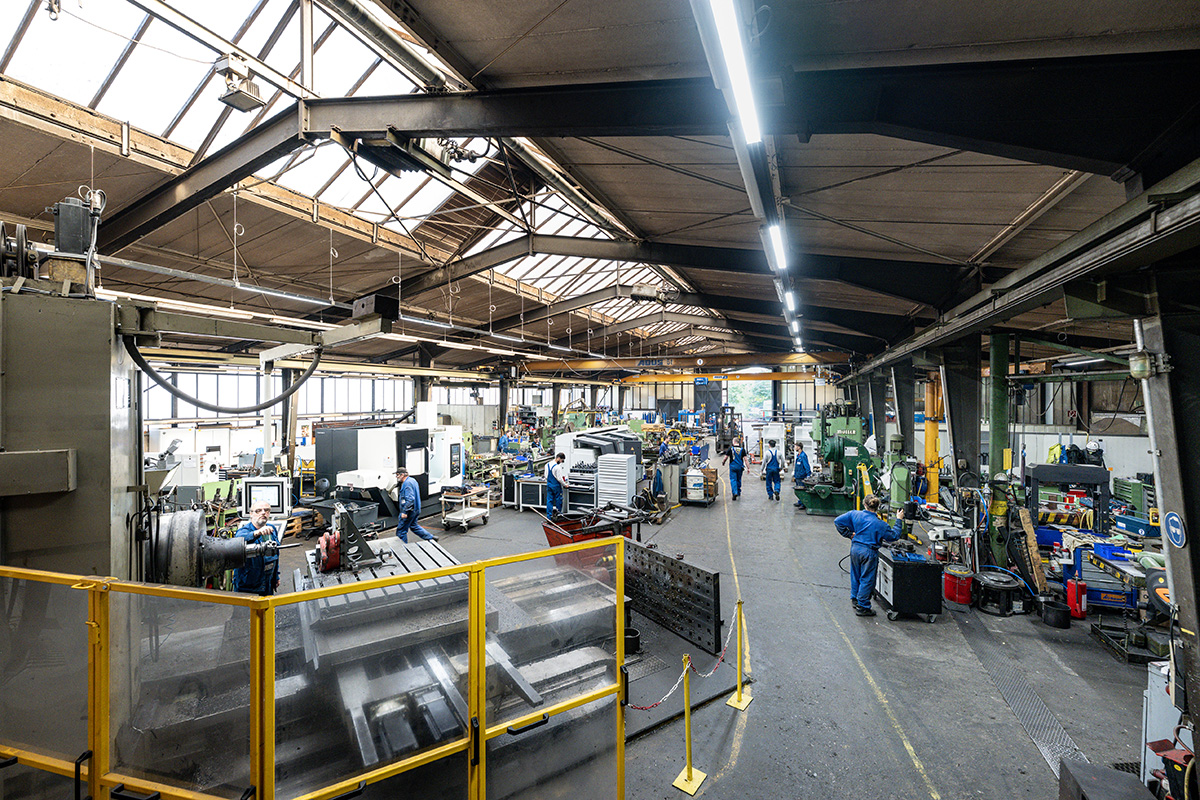 Panorama einer Werkhalle mit Maschinen, Arbeitsplätzen und Mitarbeitern in blauer Arbeitskleidung
