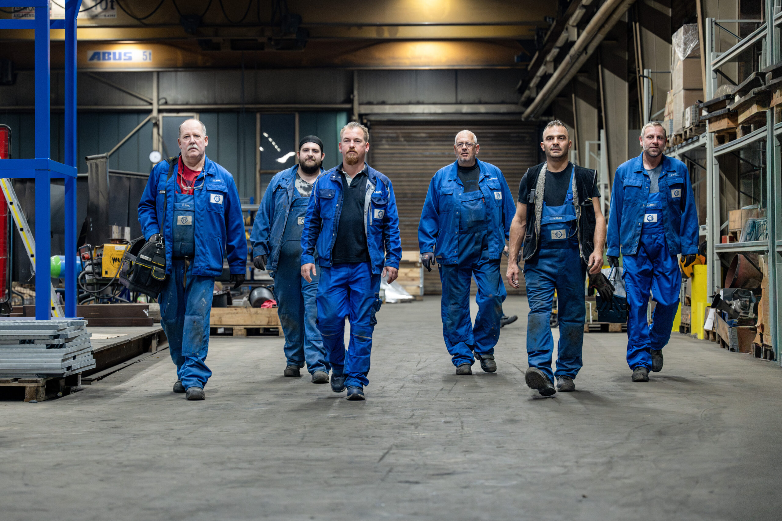 Gruppe von sechs Industriearbeitern in blauer Arbeitskleidung läuft gemeinsam durch eine Werkhalle
