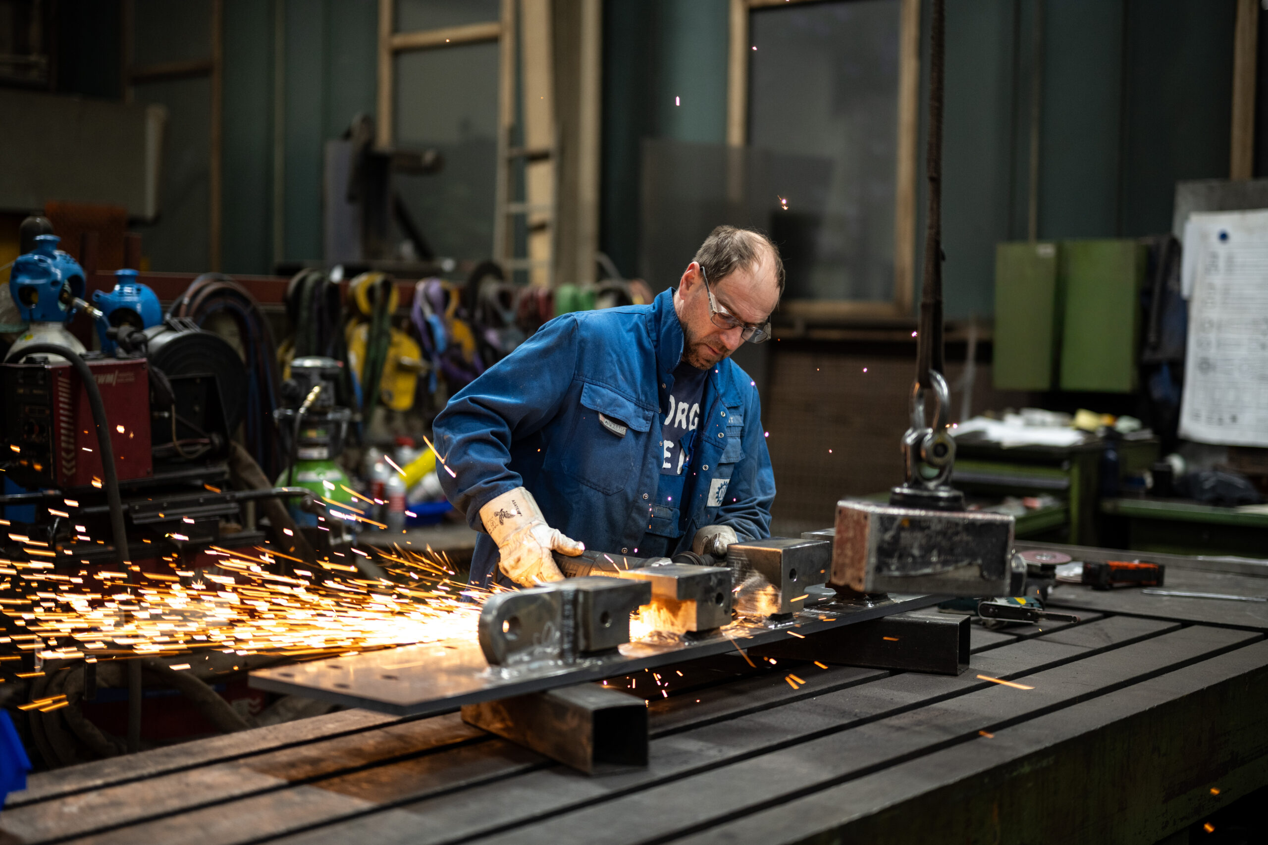 Arbeiter in blauer Schutzkleidung schleift Metallteil mit Winkelschleifer, Funken sprühen in der Werkstatt