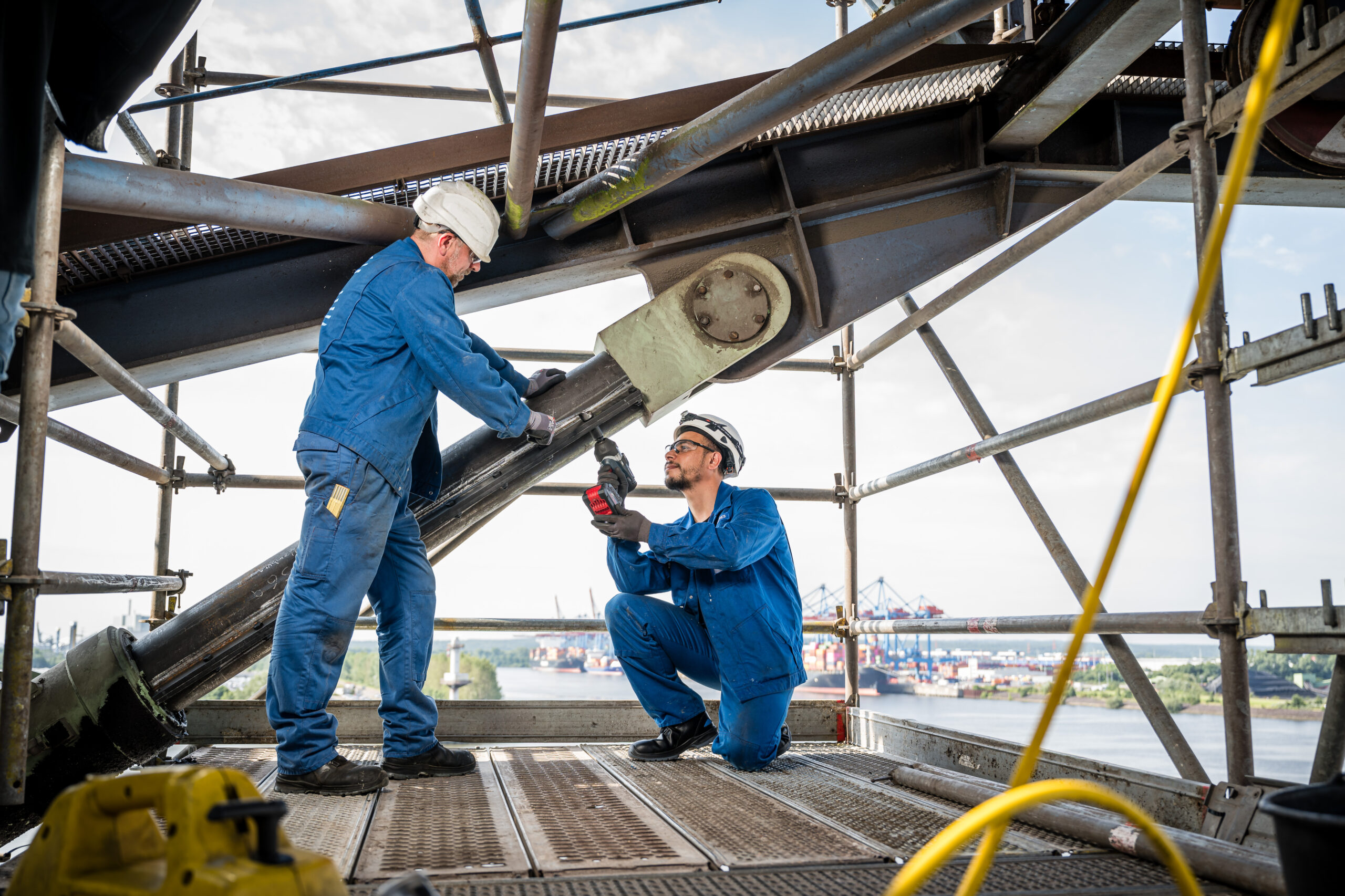 Zwei Industriearbeiter in blauer Schutzkleidung und Helm arbeiten auf einem Gerüst an einer großen Stahlkonstruktion mit Blick auf den Hafen