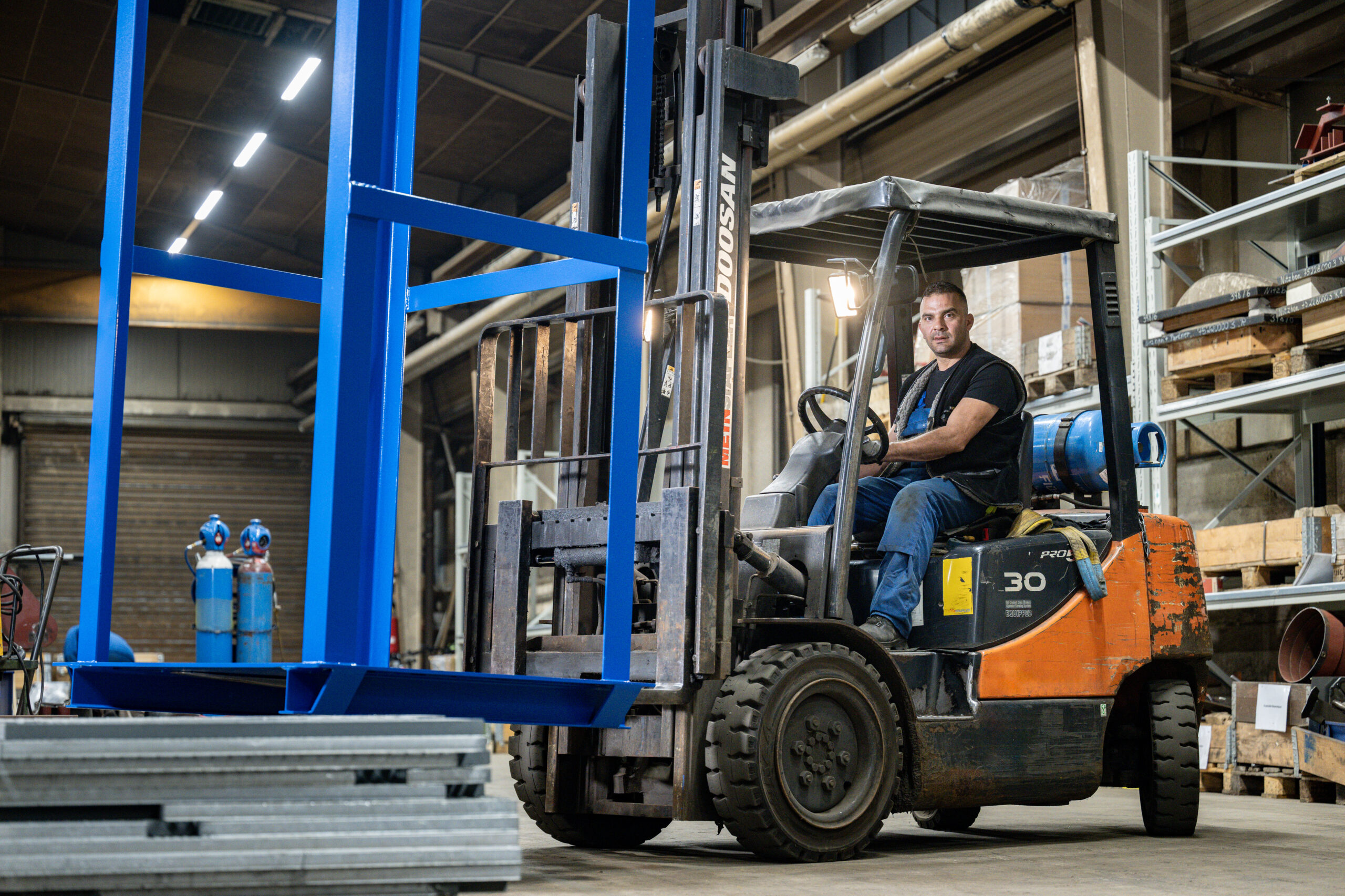 Mitarbeiter fährt mit einem Gabelstapler ein großes blaues Metallgestell in der Werkhalle