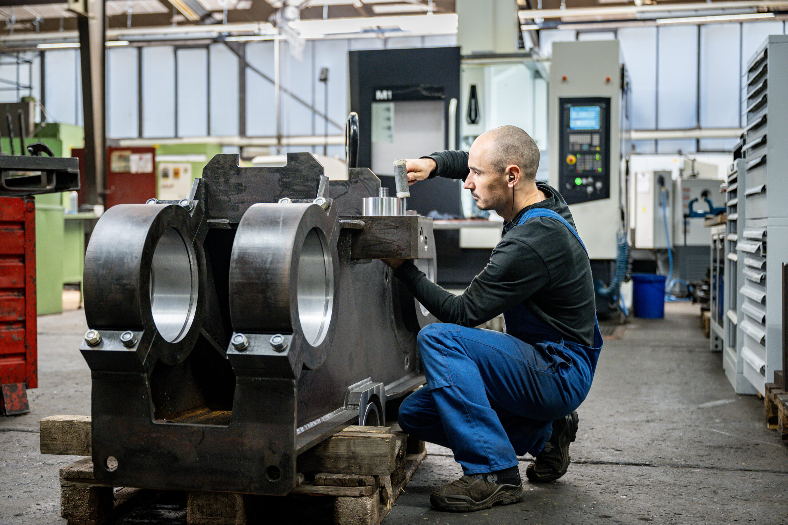 Mitarbeiter in blauer Arbeitskleidung arbeitet mit Hammer an großem Metallbauteil in der Werkhalle