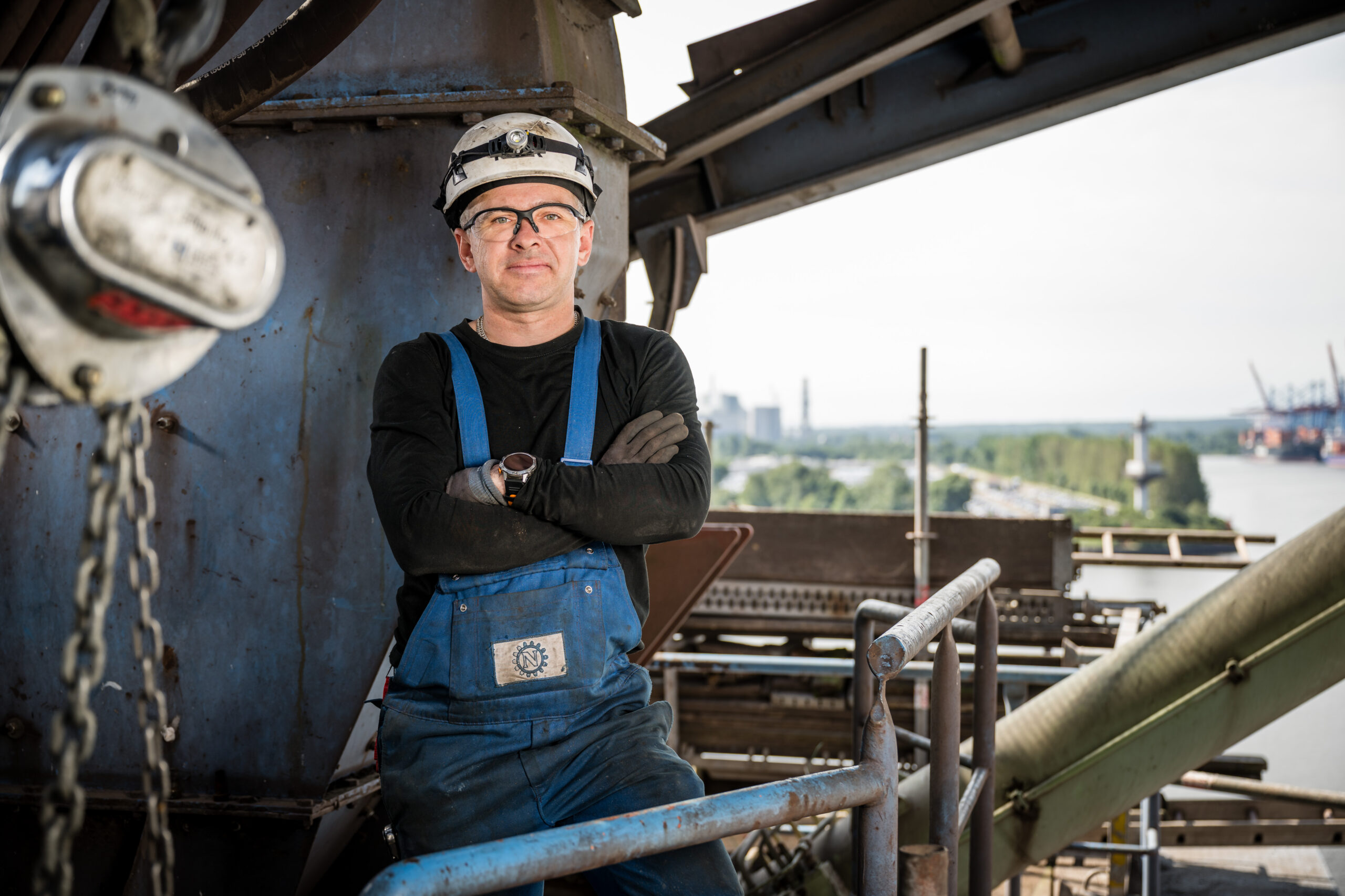 Industriearbeiter in Schutzkleidung steht mit verschränkten Armen vor Hafenanlage mit weitem Blick