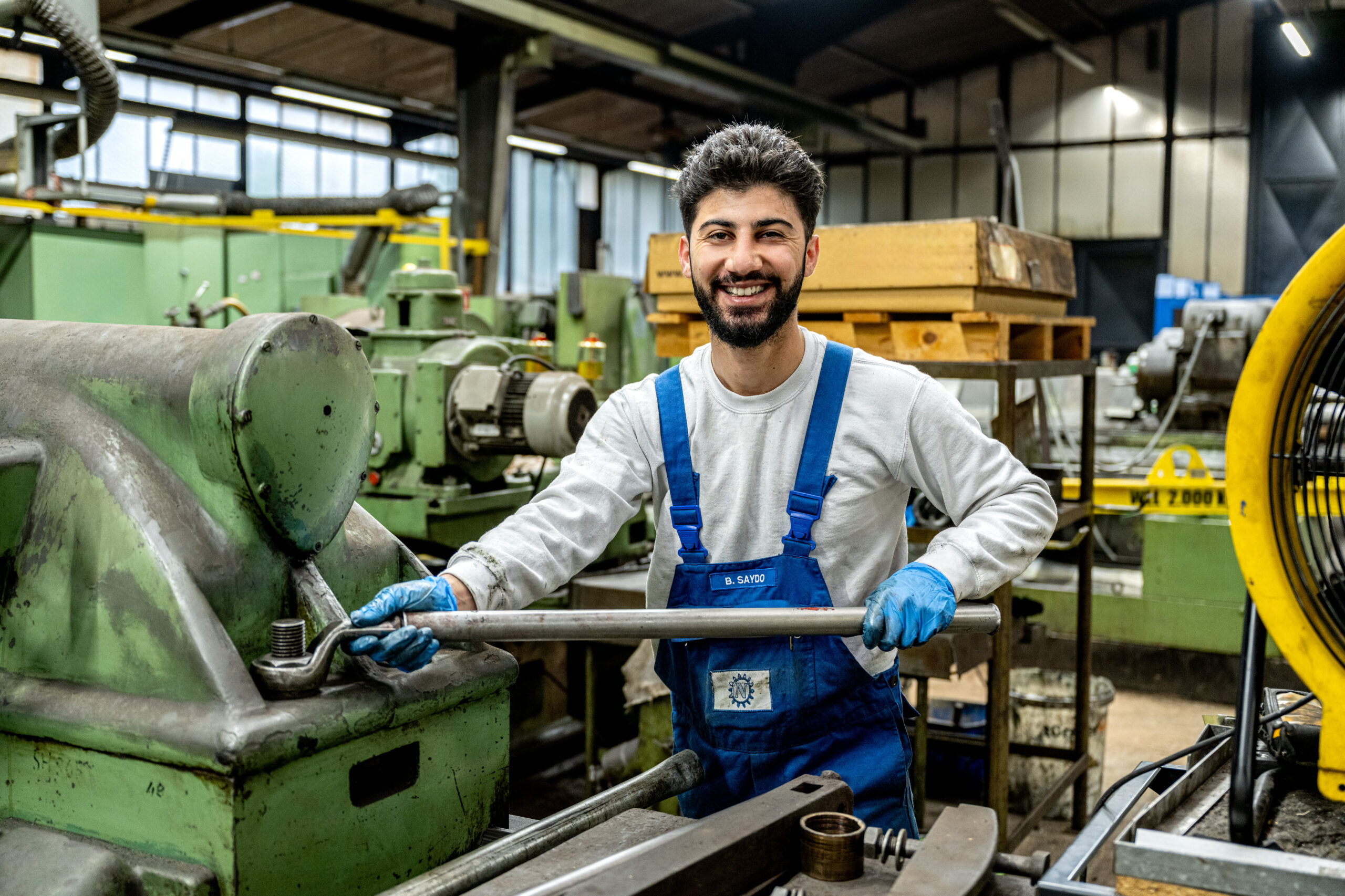 Junger Mitarbeiter in blauer Arbeitskleidung arbeitet lächelnd an einer Maschine in der Werkhalle