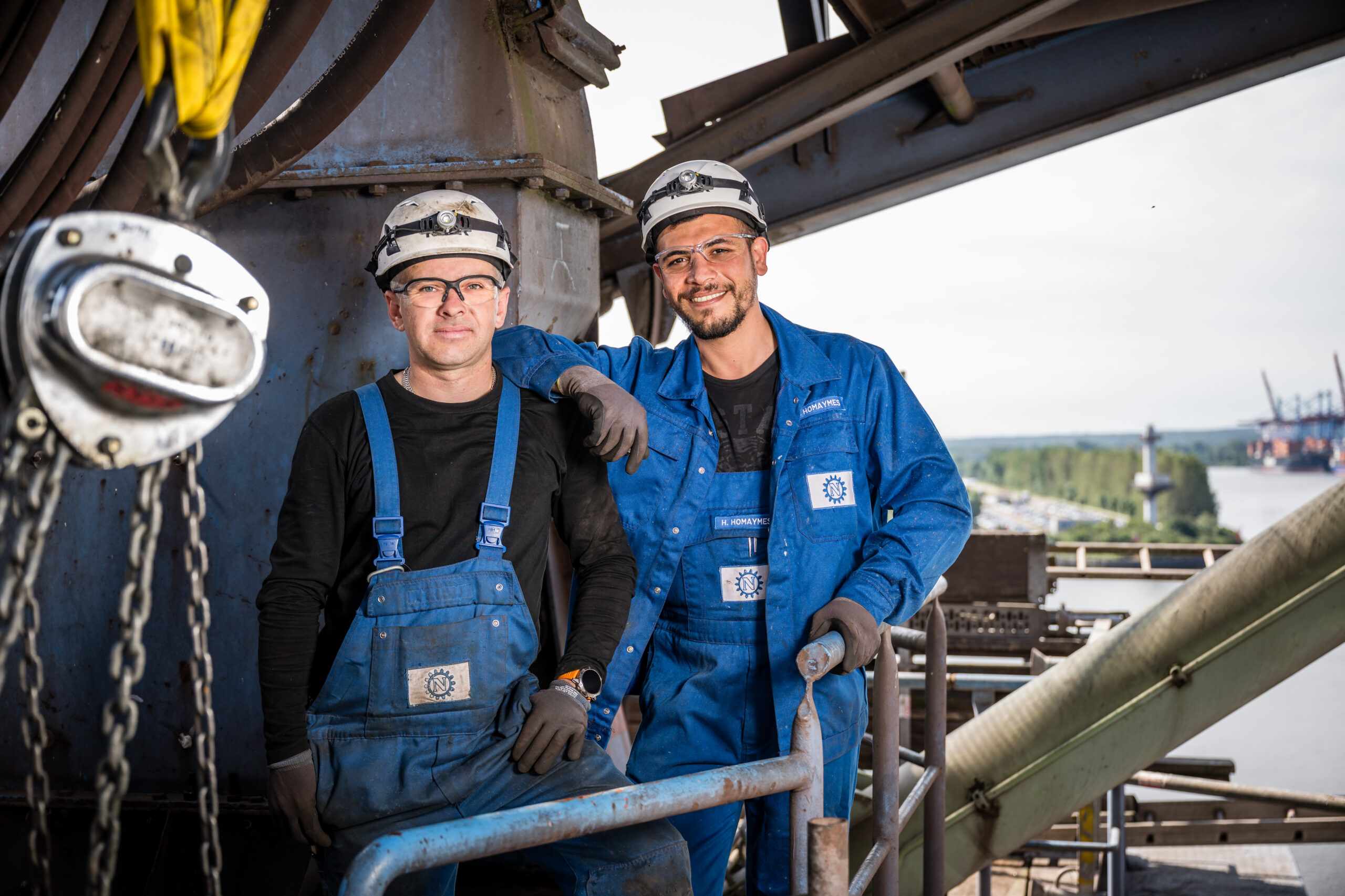 Zwei Industriearbeiter in blauen Overalls stehen lächelnd auf einer Anlage mit Blick über den Hafen