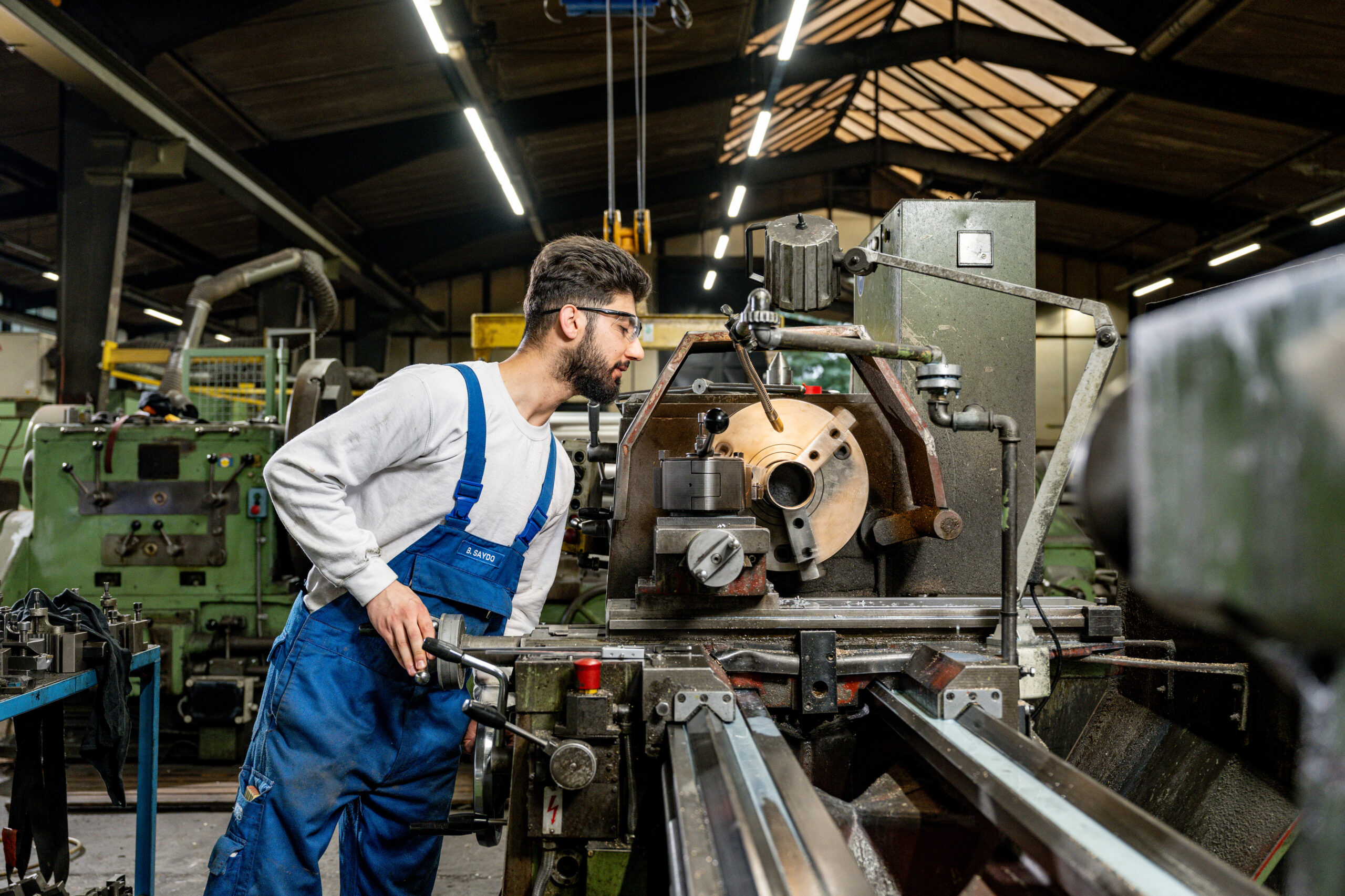 Maschinenbediener in blauer Arbeitskleidung und Schutzbrille arbeitet konzentriert an einer Drehmaschine in der Werkhalle