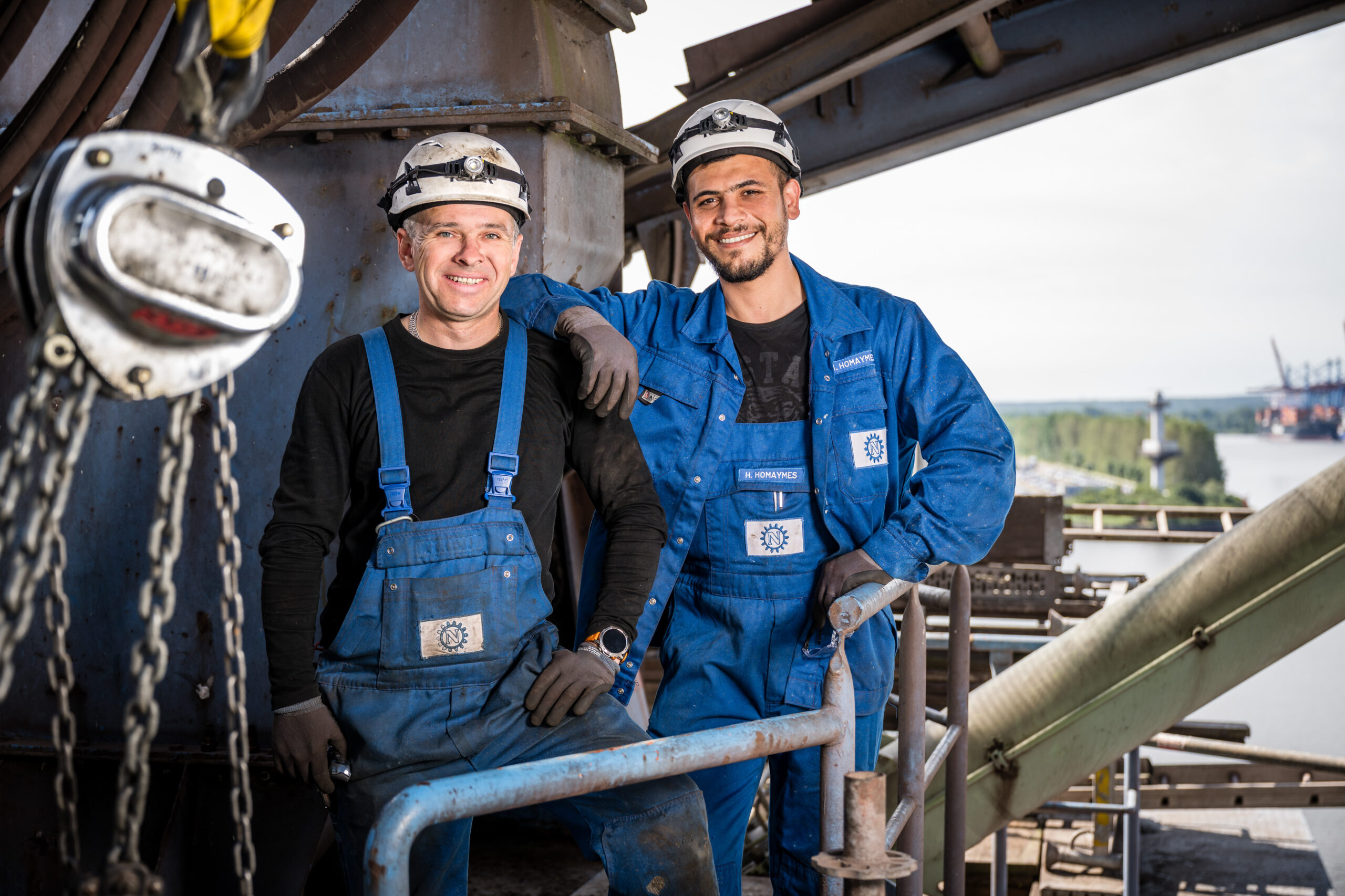 Zwei Industriearbeiter in blauen Overalls stehen lächelnd auf einer Anlage mit Blick über den Hafen