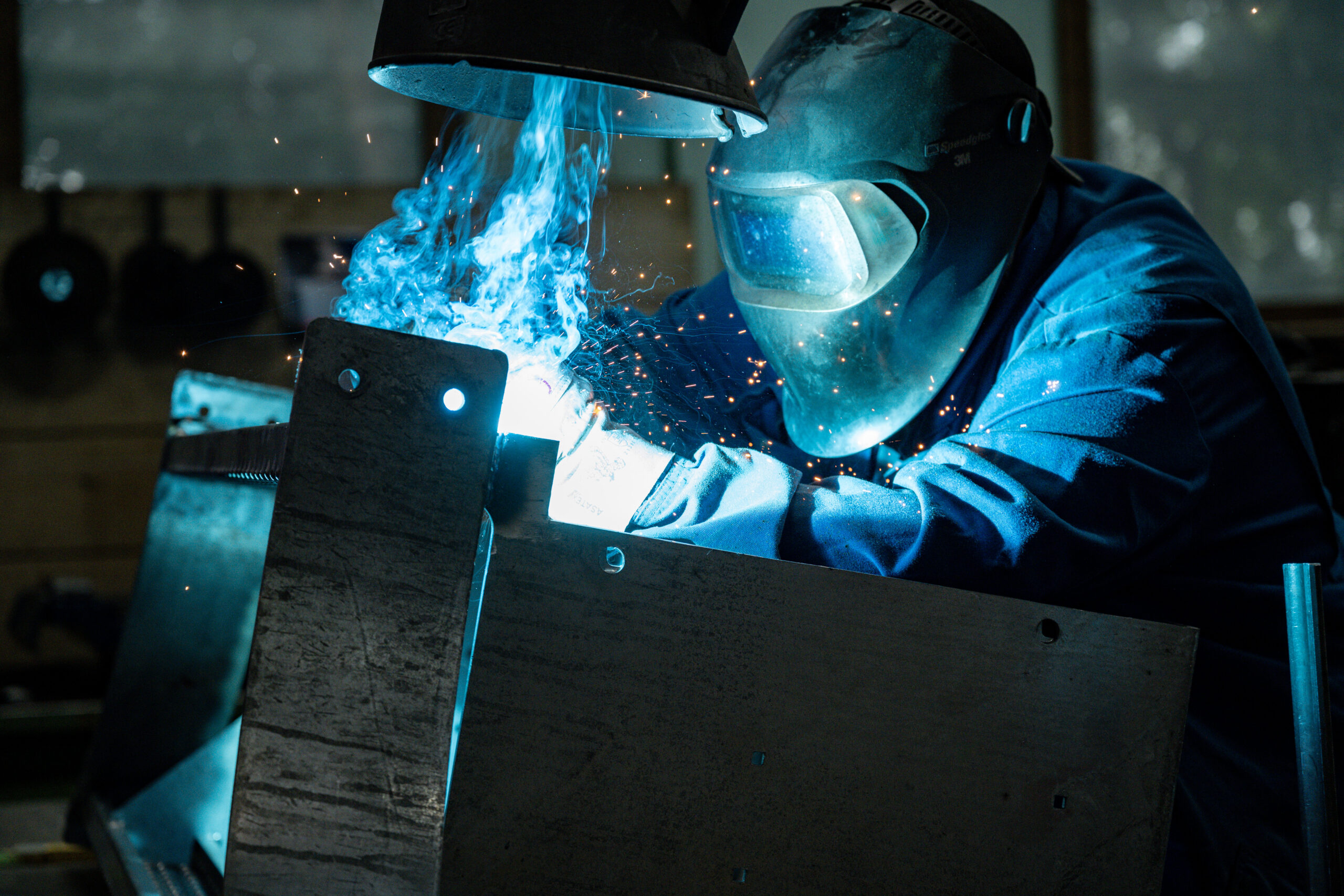 Schweißer mit Schutzhelm bearbeitet ein Metallteil, helles Schweißlicht und Funkenflug in der Werkhalle