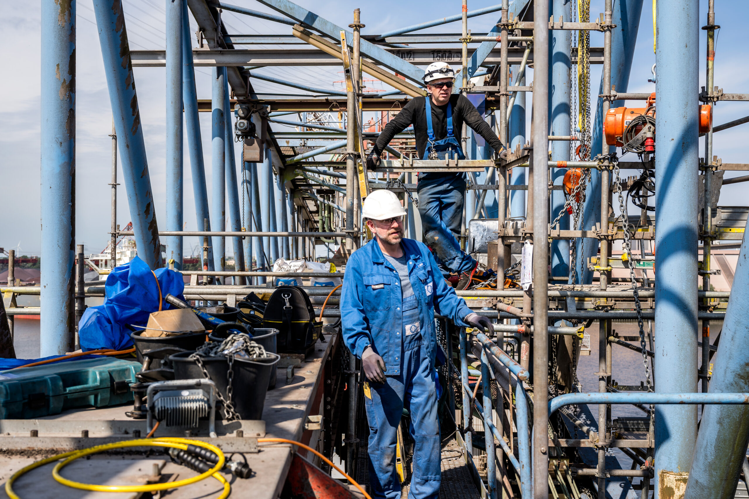 Zwei Industriearbeiter mit Schutzhelmen arbeiten auf einem Gerüst an einer großen Stahlkonstruktion über dem Wasser