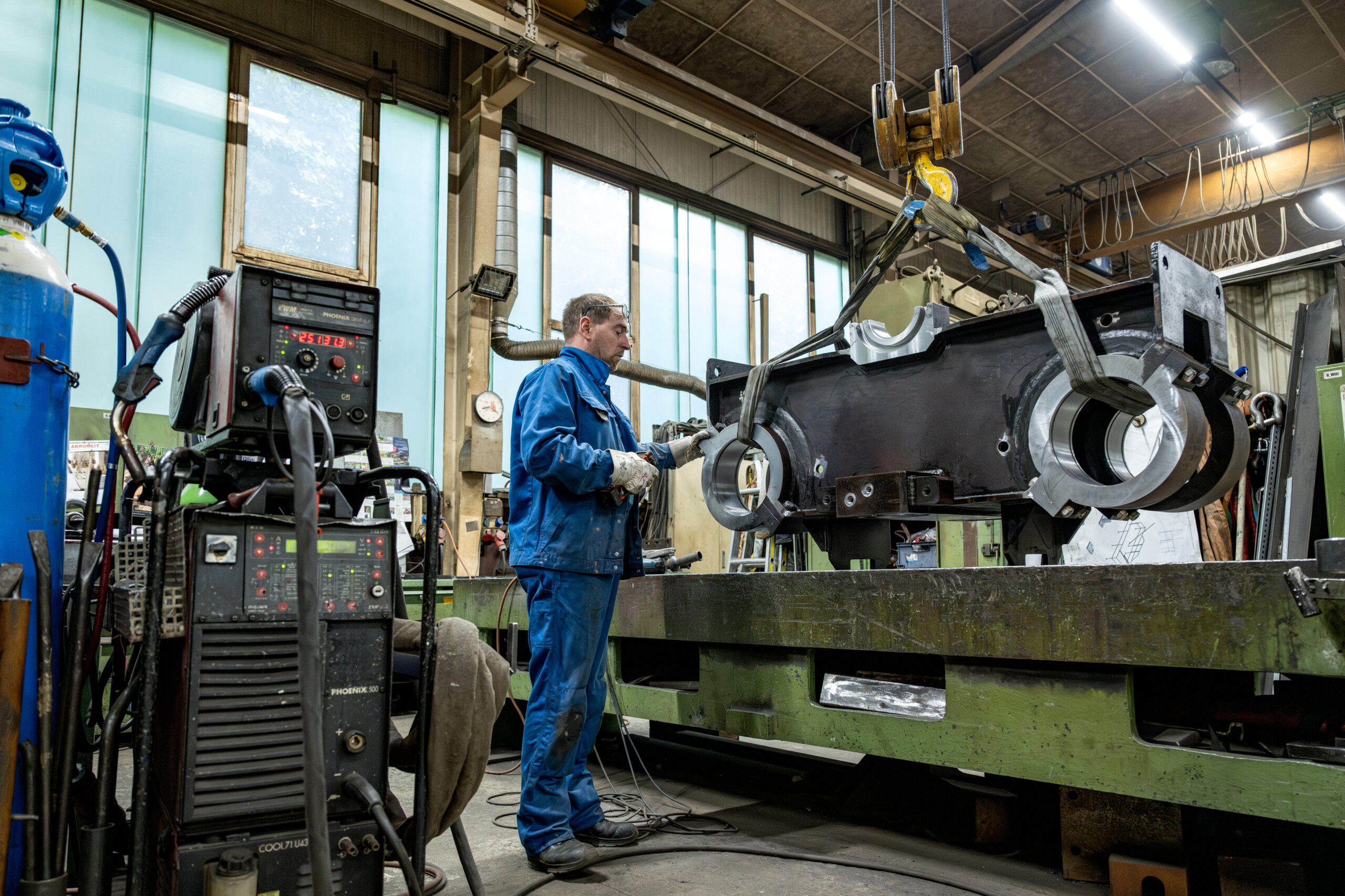 Mitarbeiter in blauer Arbeitskleidung richtet mit Kranunterstützung ein großes Metallbauteil in der Werkhalle aus