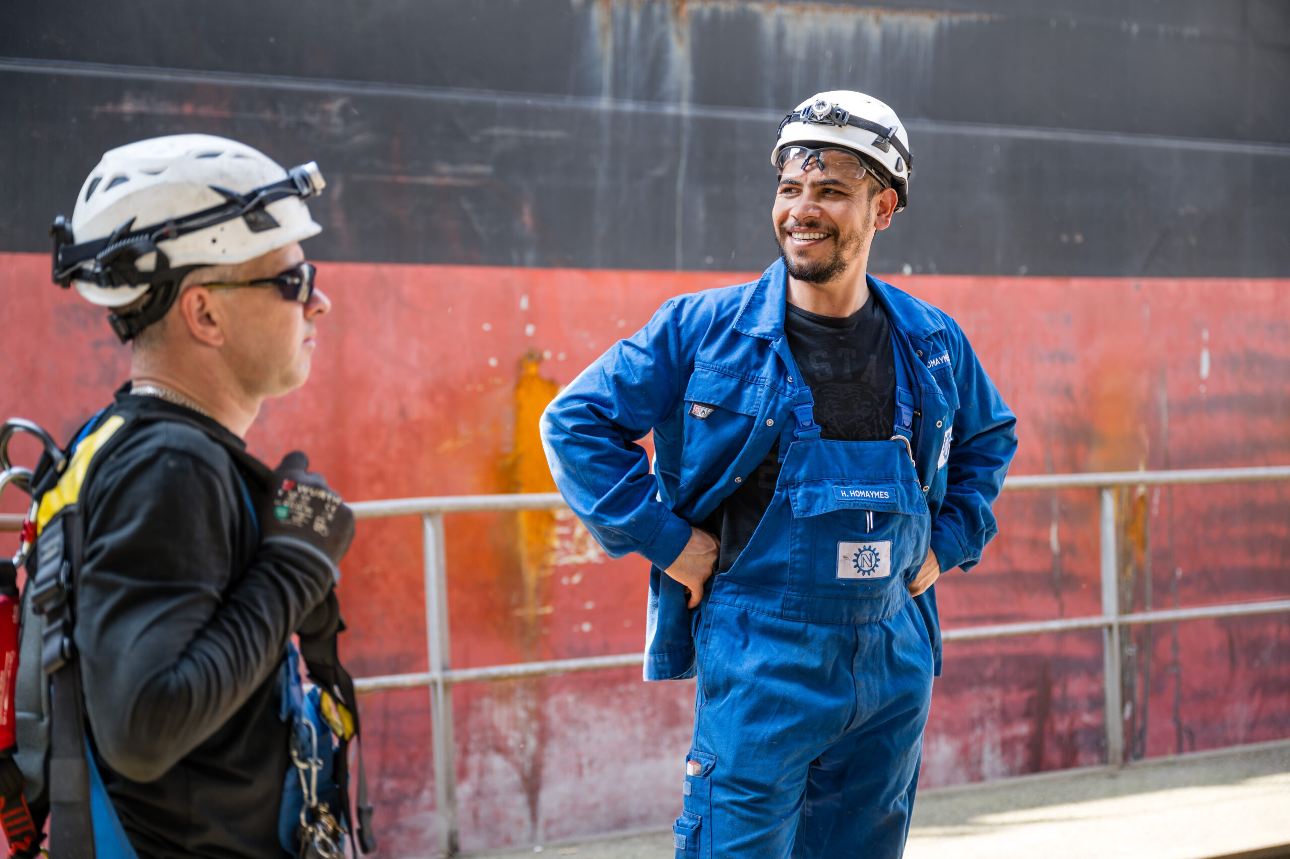 Zwei Industriearbeiter im Gespräch vor einem Frachtschiff, einer lacht herzlich
