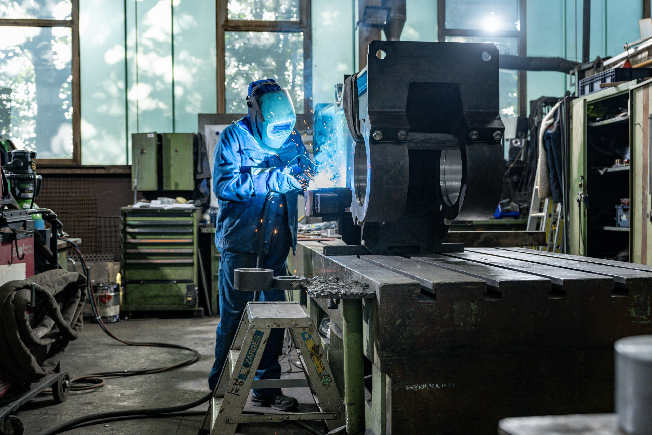 Arbeiter mit Schweißhelm bearbeitet ein großes Metallteil auf einer Werkbank, helles Schweißlicht und Funkenflug
