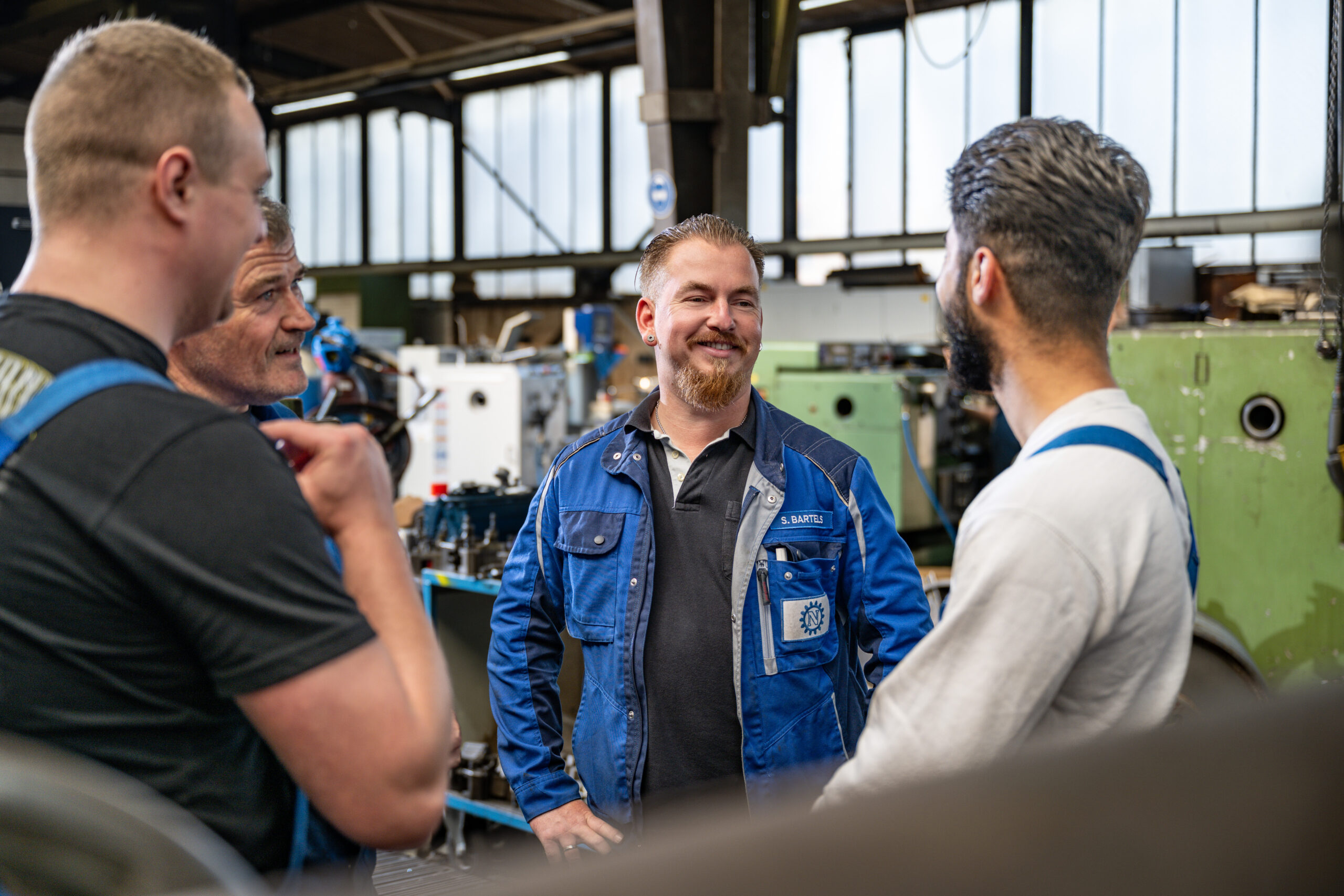 Gruppe von Mitarbeitern in blauer Arbeitskleidung unterhält sich lachend in einer Werkhalle