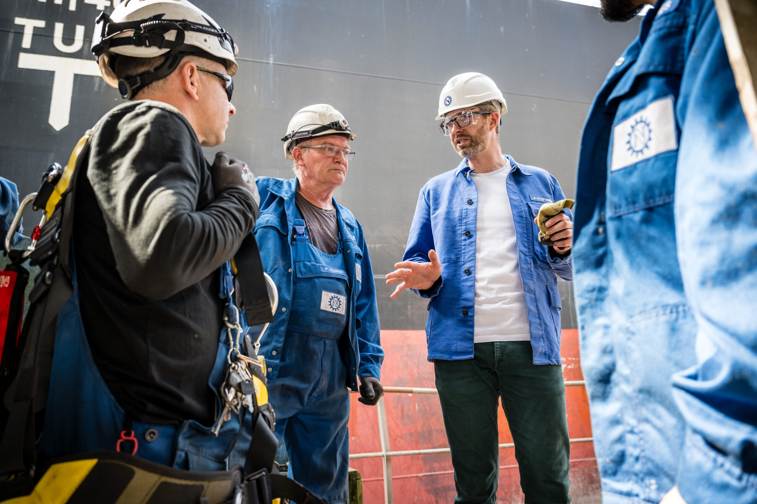 Gruppe von Mitarbeitern mit Schutzhelmen bespricht Arbeitsschritte in einer Industrieanlage
