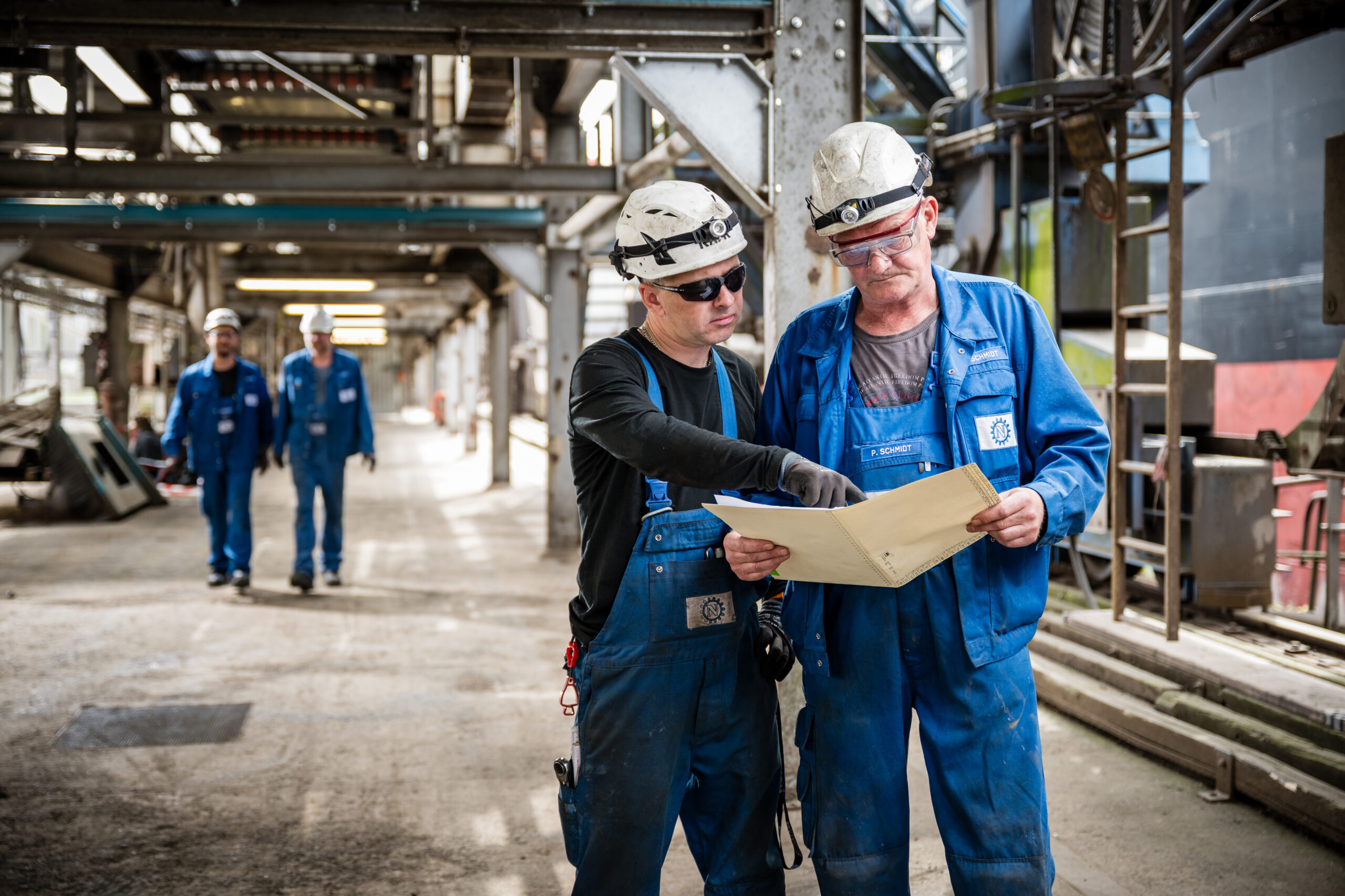 Zwei Mitarbeiter in blauer Arbeitskleidung und Helmen prüfen gemeinsam Unterlagen in einer Industrieanlage
