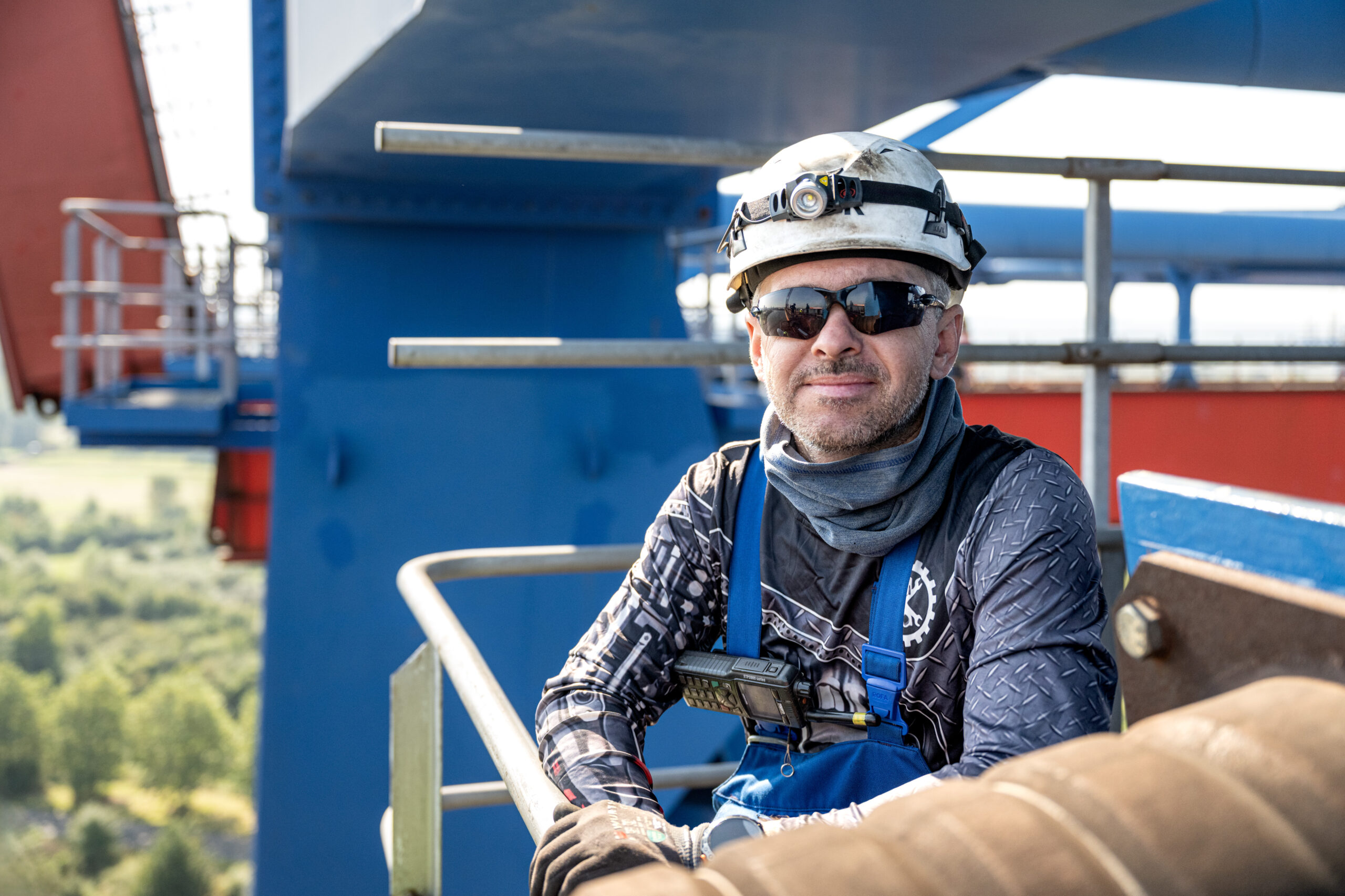 Arbeiter mit Schutzhelm, Handschuhen und Sicherheitsgeschirr sitzt auf einer Plattform einer Stahlkonstruktion