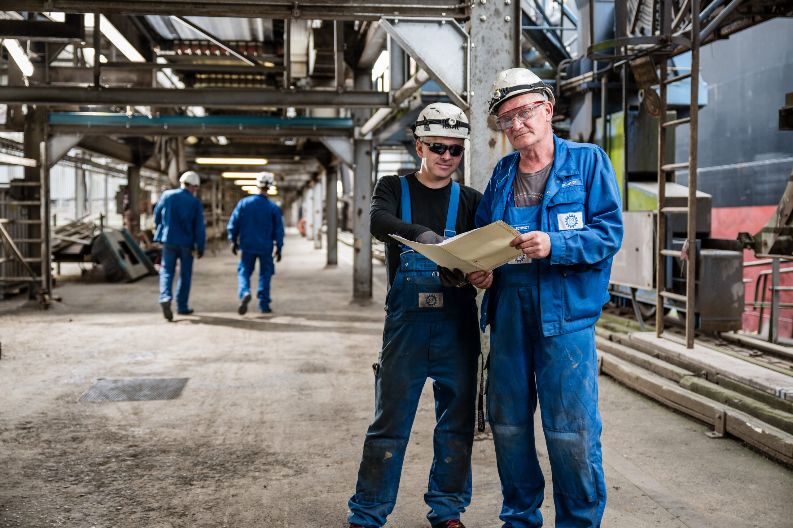 Zwei Mitarbeiter in blauer Arbeitskleidung und Helmen prüfen gemeinsam Unterlagen in einer Industrieanlage