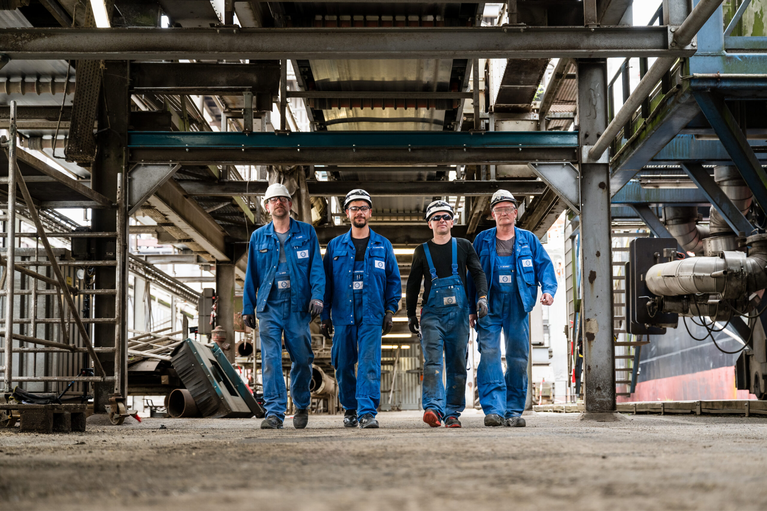 Vier Mitarbeiter in blauer Arbeitskleidung und Helmen laufen selbstbewusst durch eine Industrieanlage