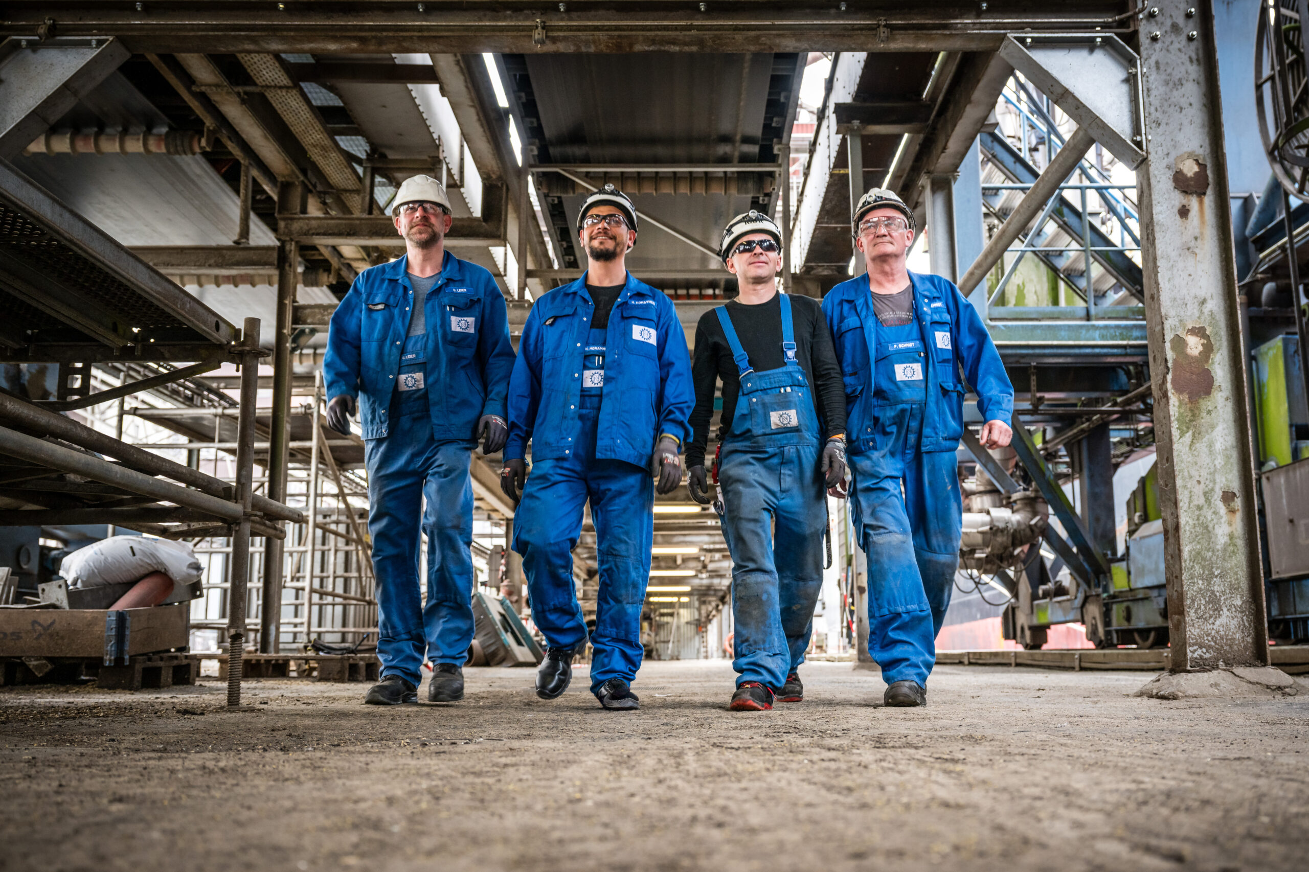 Vier Mitarbeiter in blauer Arbeitskleidung und Helmen laufen selbstbewusst durch eine Industrieanlage