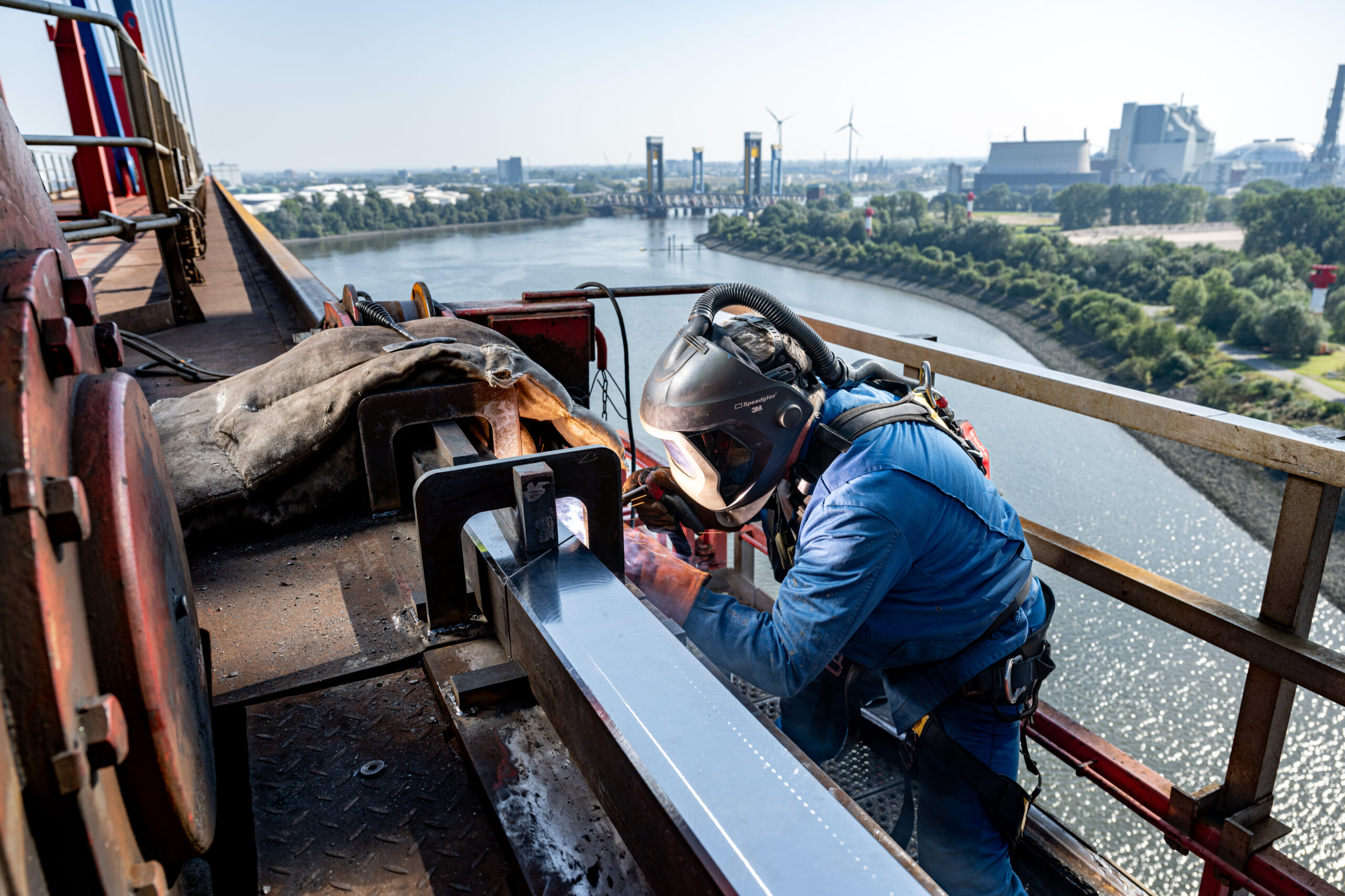 Arbeiter mit Schutzhelm und Sicherheitsgeschirr schweißt an einer Stahlkonstruktion hoch über einem Fluss