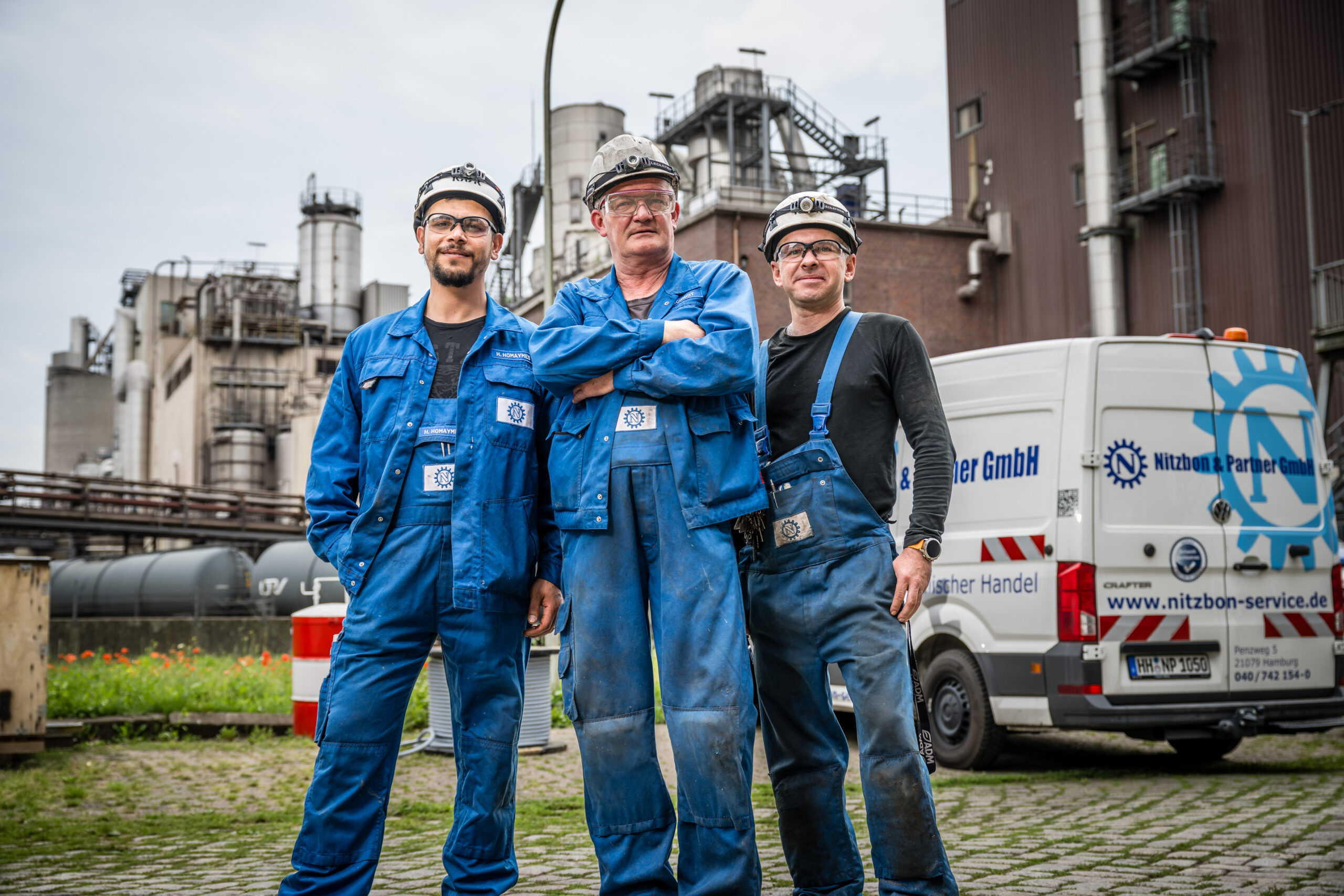 Drei Mitarbeiter in blauer Arbeitskleidung mit Helmen stehen selbstbewusst vor Industrieanlage und Servicefahrzeug