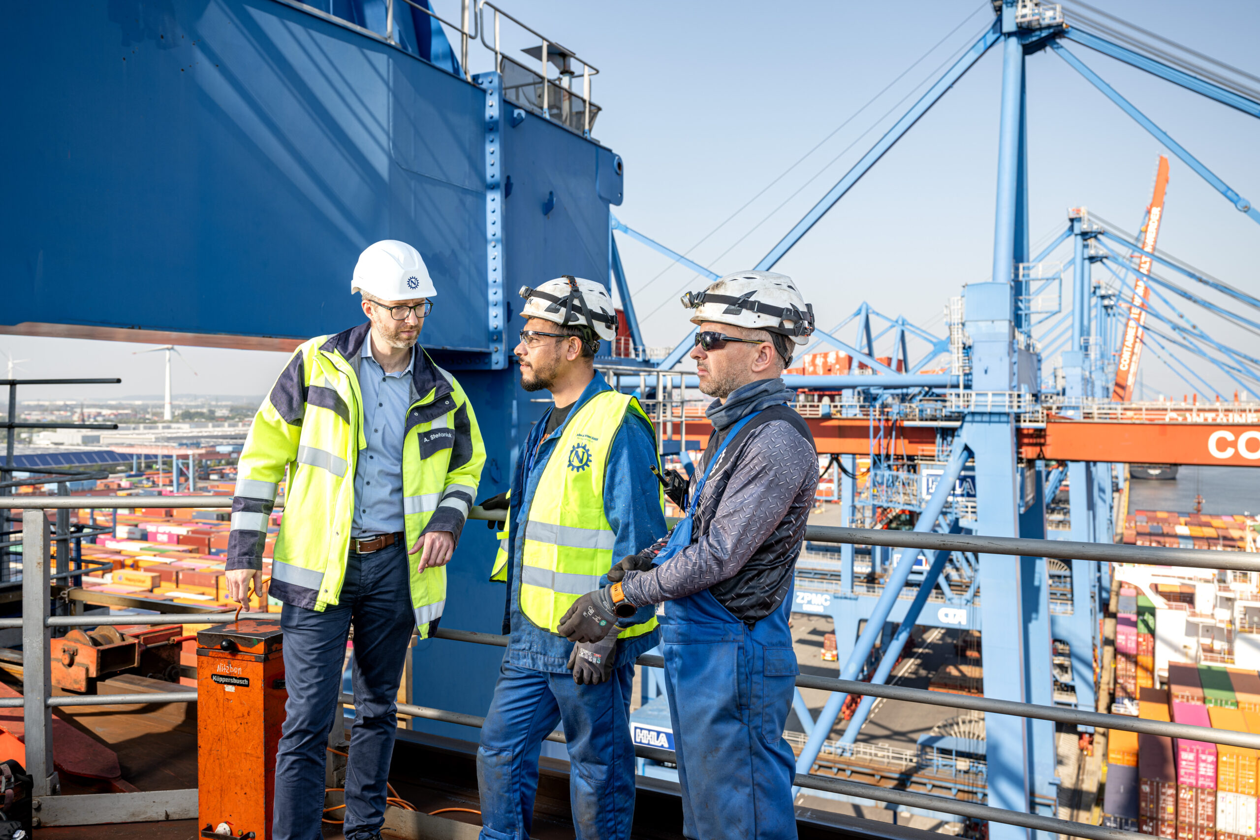 Drei Industriearbeiter mit Schutzhelmen und Warnwesten besprechen sich auf einer Hafenanlage vor großen Containerkränen