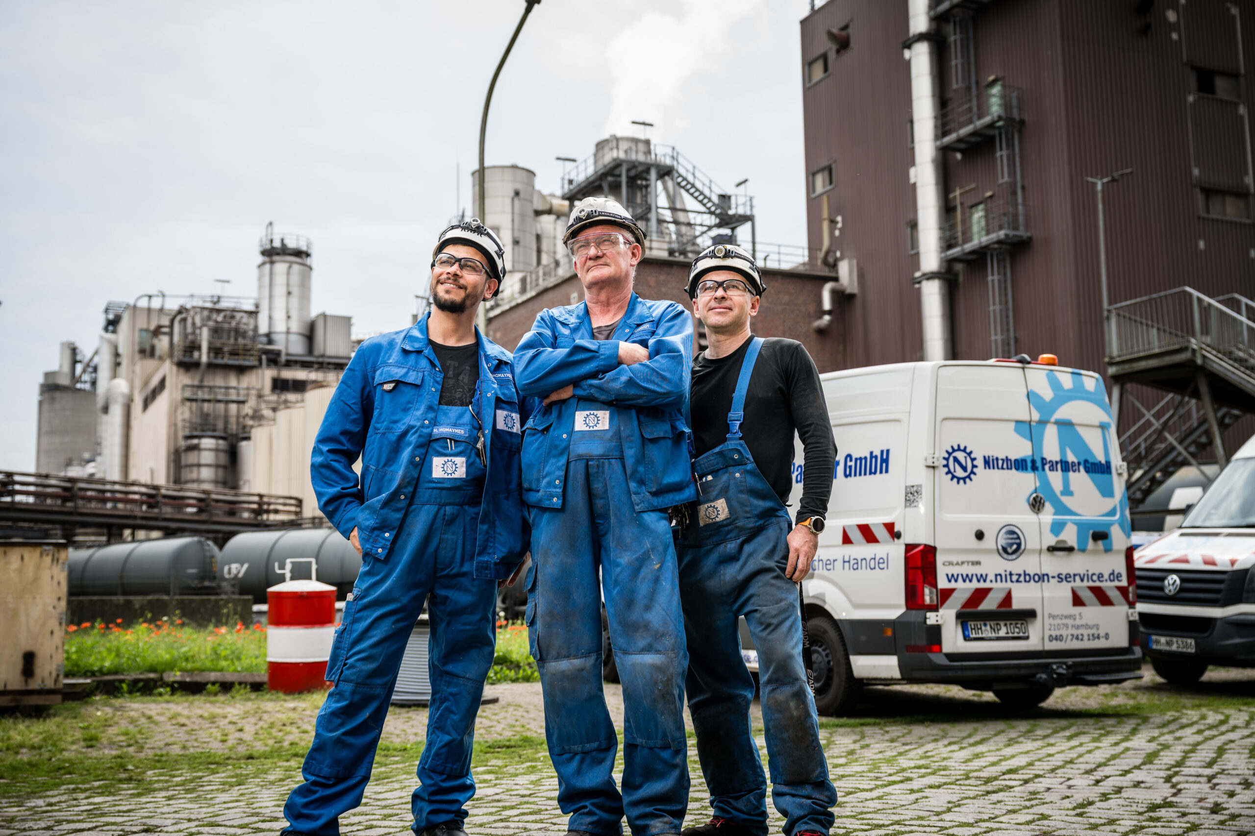 Drei Mitarbeiter in blauer Arbeitskleidung mit Helmen stehen selbstbewusst vor Industrieanlage und Servicefahrzeug