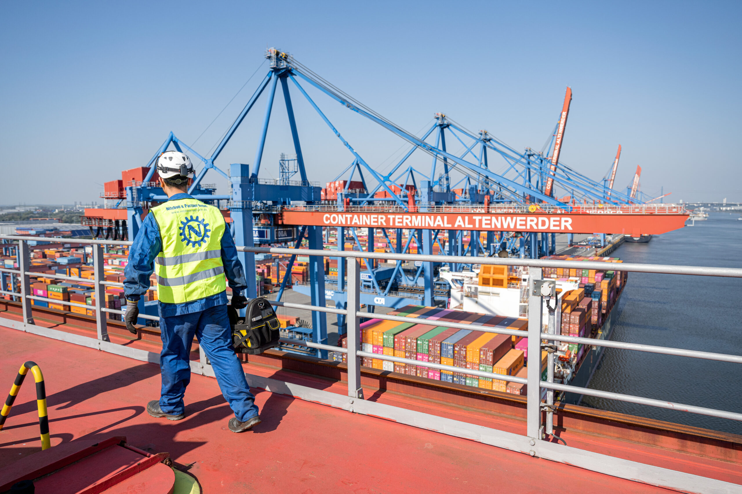 Arbeiter in Schutzkleidung mit Werkzeugtasche blickt auf Containerterminal Altenwerder mit Kränen und Containerschiffen