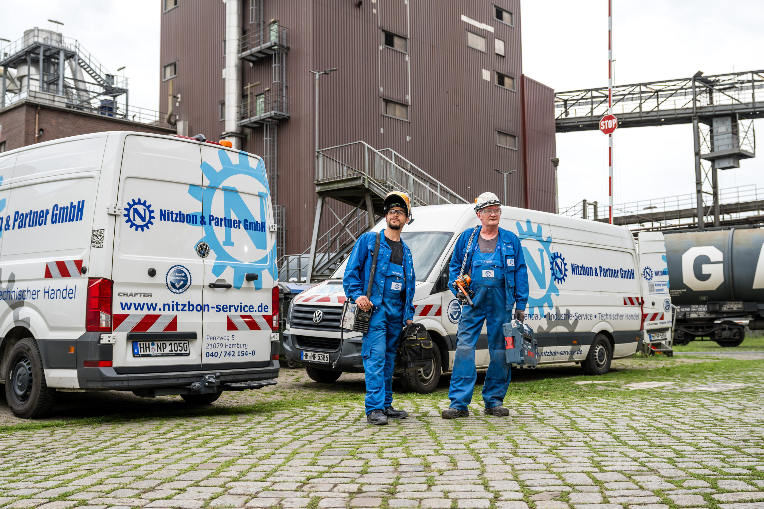 Zwei Mitarbeiter in blauer Arbeitskleidung mit Werkzeugtaschen gehen vor Service-Fahrzeugen von Nitzbon & Partner GmbH