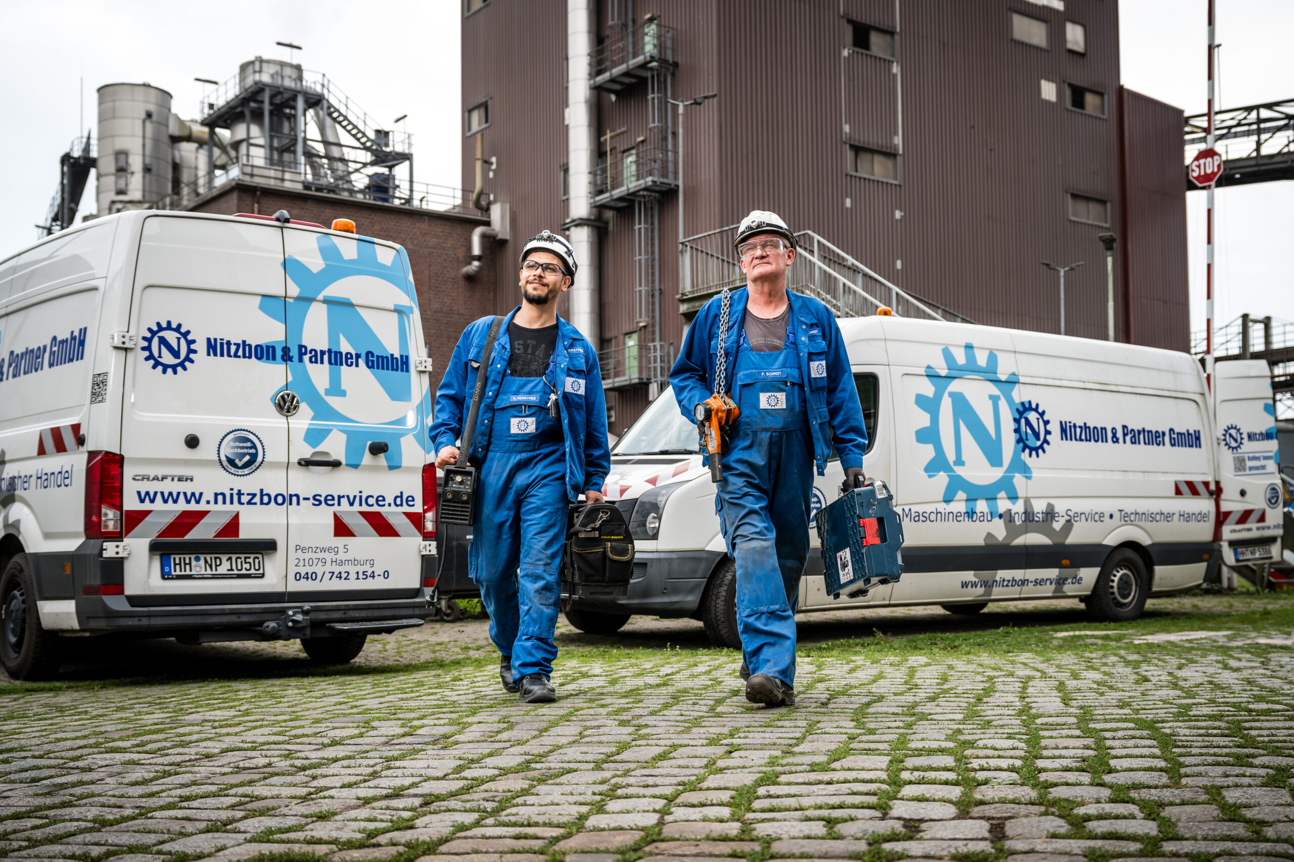 Zwei Mitarbeiter in blauer Arbeitskleidung mit Werkzeugtaschen gehen vor Service-Fahrzeugen von Nitzbon & Partner GmbH